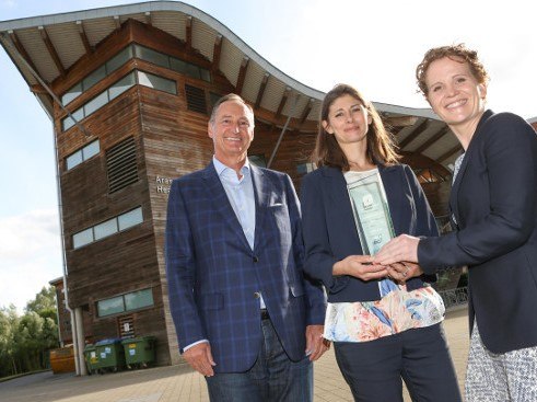 A man in blue checkered suit stands with two women holding a prize in front of a modern building.