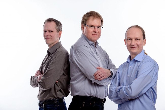 Three men in grey and blue shirts stand with their arms folded.