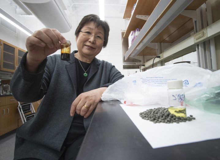 A woman sits at a lab bench beside a small mound of pellets, a container of gasoline-like fuel and a plastic bag. She holds a small container of dark oil produced through recycling plastic.