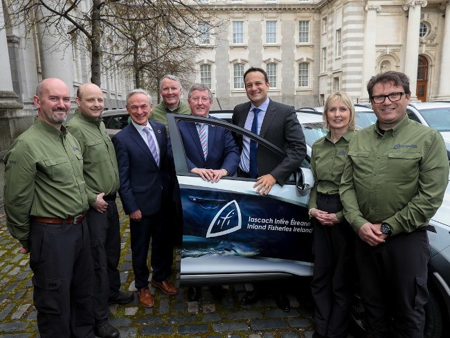 Seven men and one woman stand beside a new electric vehicle outside Irish government buildings.