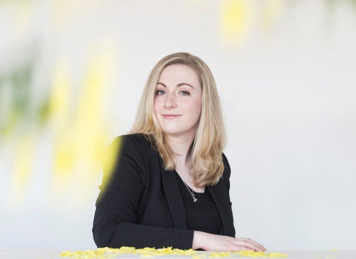 Woman with blonde hair and black jacket sits with one arm on a table strewn with flower petals.