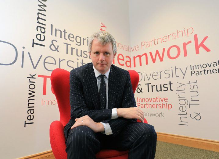 Man in pin-striped suit sitting in red chair stares intensely at camera.