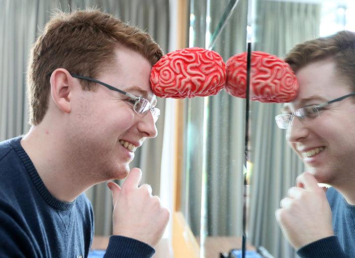 James Blackwell leans into his reflection in a mirror, propping his head up against a small model of the brain.