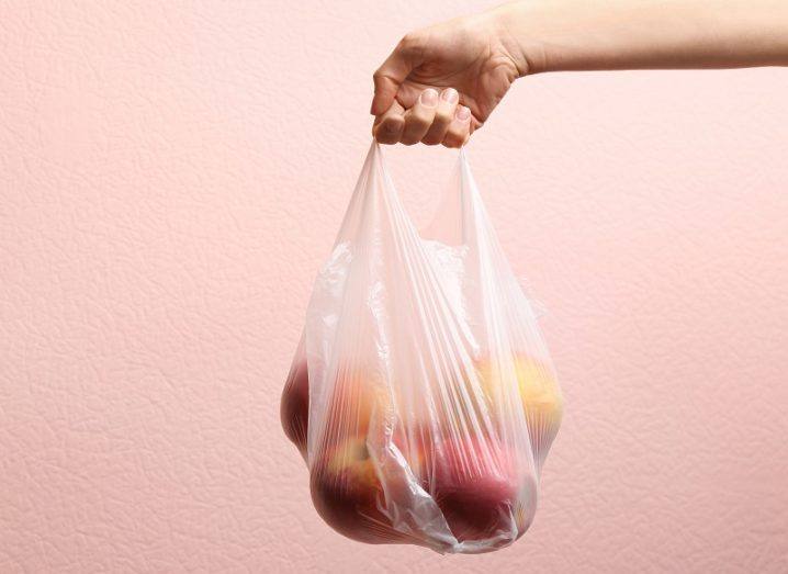 Close-up of a woman holding a plastic bag with red apples against a pink background.