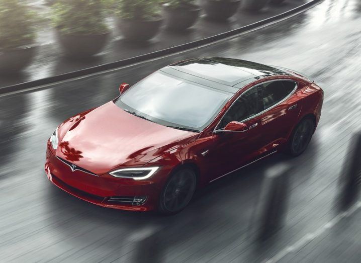 A red Tesla Model S driving on a wet road with concrete and plant bollards either side.