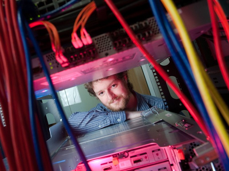 Man with brown hair and beard pokes his head through a server rack.