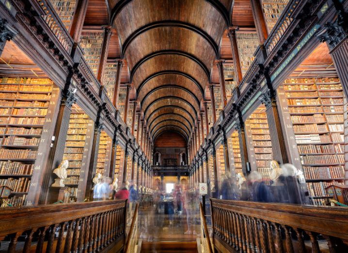 Inside the iconic Trinity College library of Dublin with people moving around the various aisles.