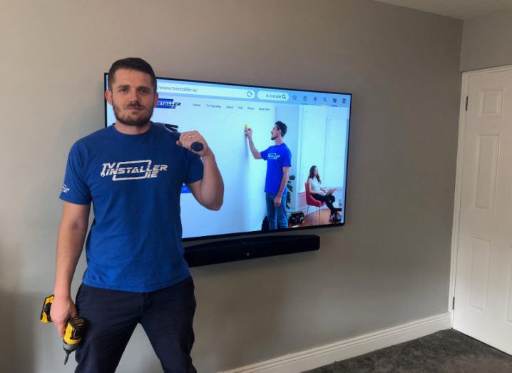 Man in blue t-shirt holding a power drill beside a newly installed flatscreen TV.