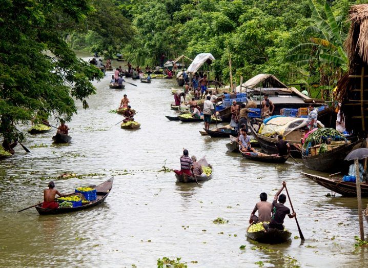 People in small boats on a river surrounded by vegetation and market stalls.