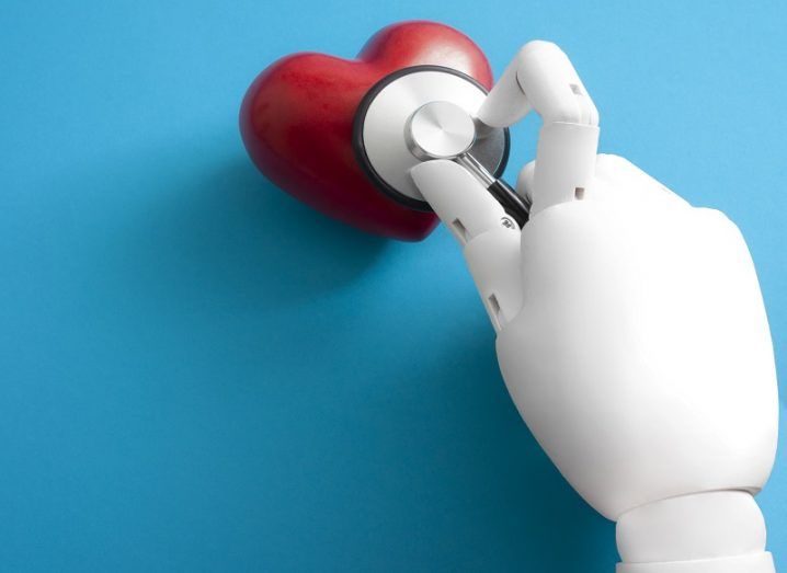 A white robotic hand holding a stethoscope to a red heart against a blue background.