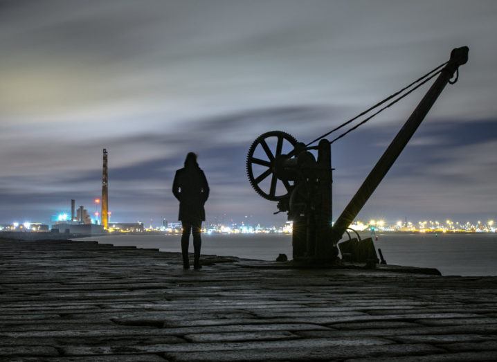 Woman stands looking at lights of Dublin beside a crane at Poolbeg light tower.