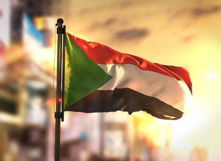 View of the flag of Sudan billowing in the breeze amid a sunset-drenched blurred backdrop possibly of a city.