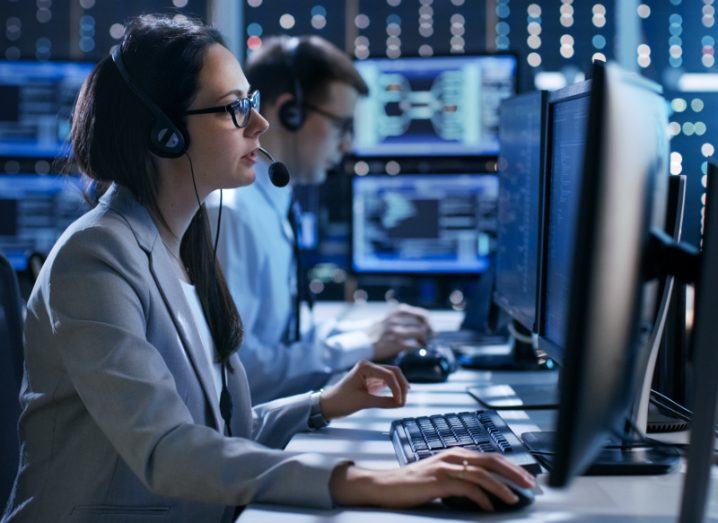 Woman in lab coat sitting at desk looking at computer and wearing headphones.