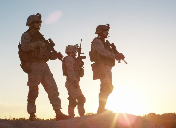 View of armed soldiers standing on an arid desert peak looking ahead of them against the backdrop of sunrise.