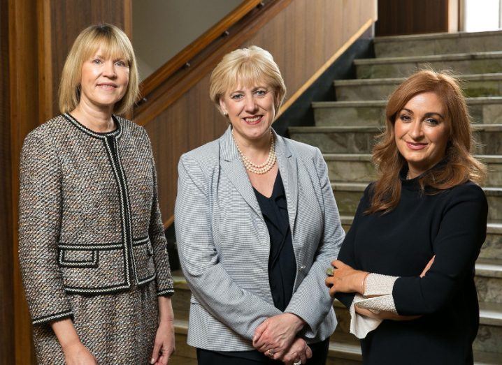 Three women in business attire pose for a photo at the bottom of a staircase.