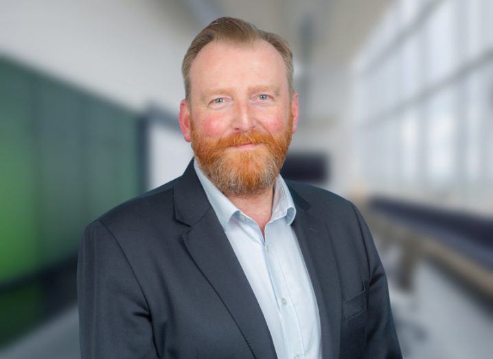 Man with red hair and red beard wearing a navy jacket and blue shirt.