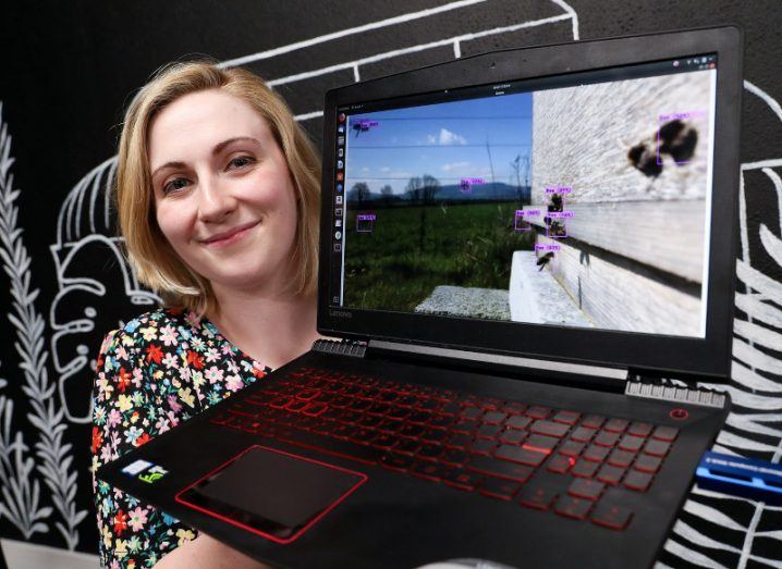 A blonde woman in a floral dress holds up a laptop displaying footage of bees being identified with markers via machine vision technology.