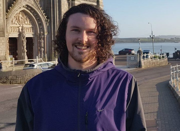 Shane McDonagh smiling and wearing a purple jacket with a church and sea view in the background.