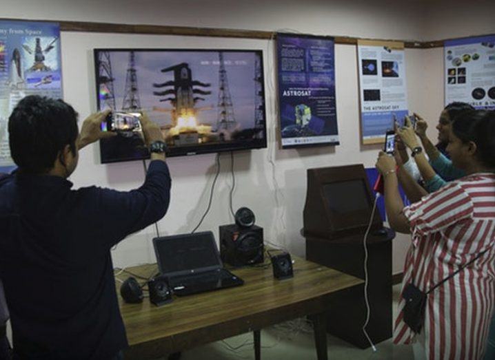 People stand around a TV screen showing the spacecraft launch, holding up their phones to capture pictures and videos.