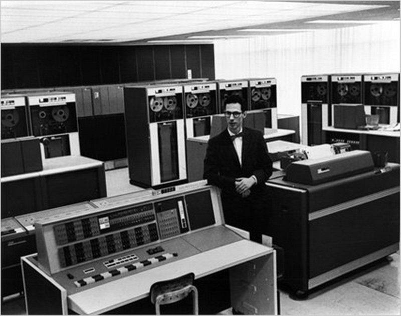 Black and white image of Fernando Carbató standing beside a number of old computers in a lab.