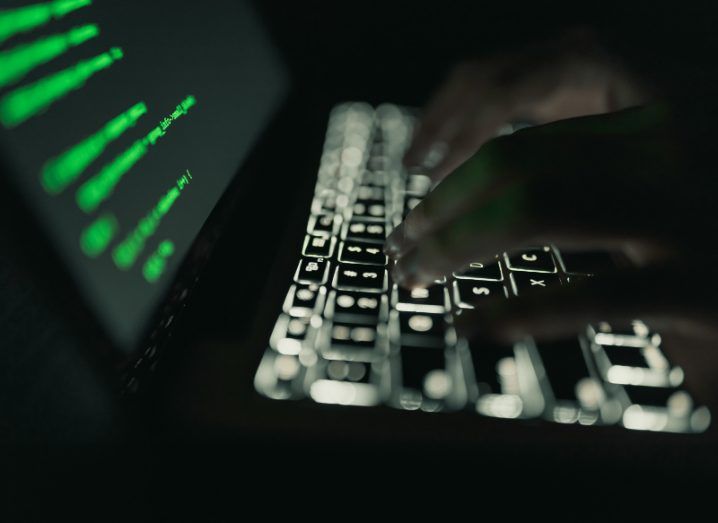 View of illuminated laptop keyboard and the silhouette of hands typing on it. The laptop screen is filled with code in green font.