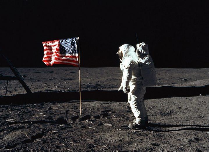 Buzz Aldrin in his spacesuit posing beside the US flag on the moon against the blackness of space.