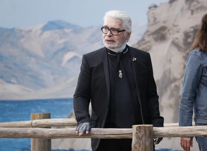 Well-dressed man with white hair in black clothes overlooking a lake from a pier.