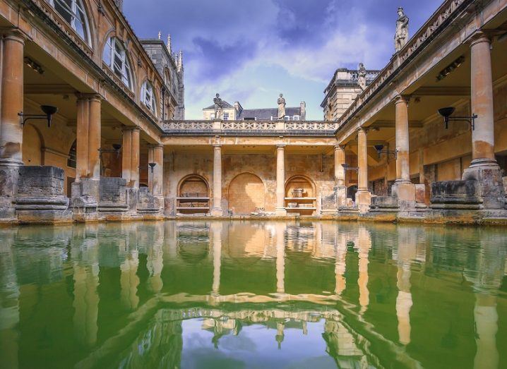 Low angled shot of green water in the square of a preserved Roman bath house with a blue sky above.