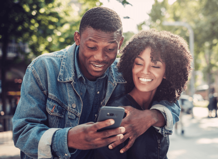 A man wearing a denim jacket has his arm around a woman in a black jumper. They are on a public street, laughing together as they both look at his mobile phone.