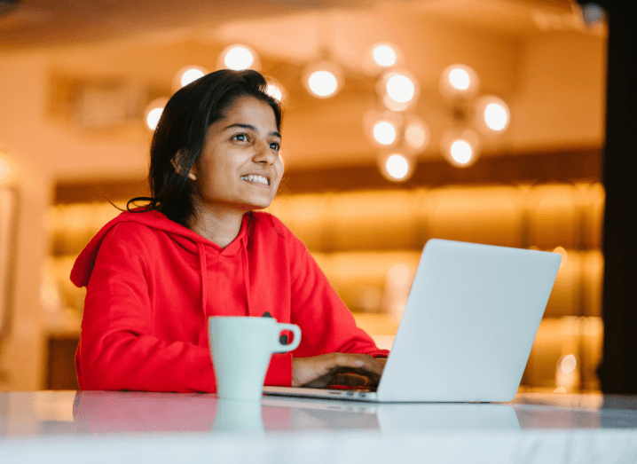 A woman in a red hoodie with dark hair, browsing the internet on a white laptop in a café.