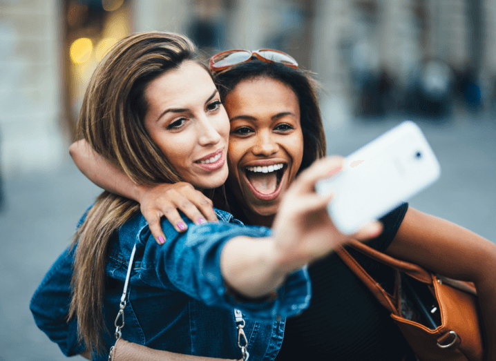 A woman in a denim jacket with dark hair holds her phone in the air to take a selfie with her friend in a black T-shirt, who is holding an orange handbag.