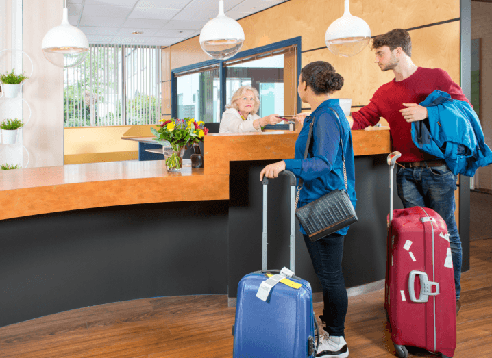 A woman in a blue shirt and black skinny jeans, checks into a hotel with a man wearing a red jumper and blue jeans. They both have luggage with them.