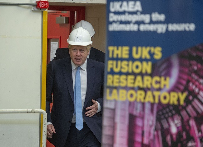 Boris Johnson in a white hardhat walking out a door partly obscured by a poster.