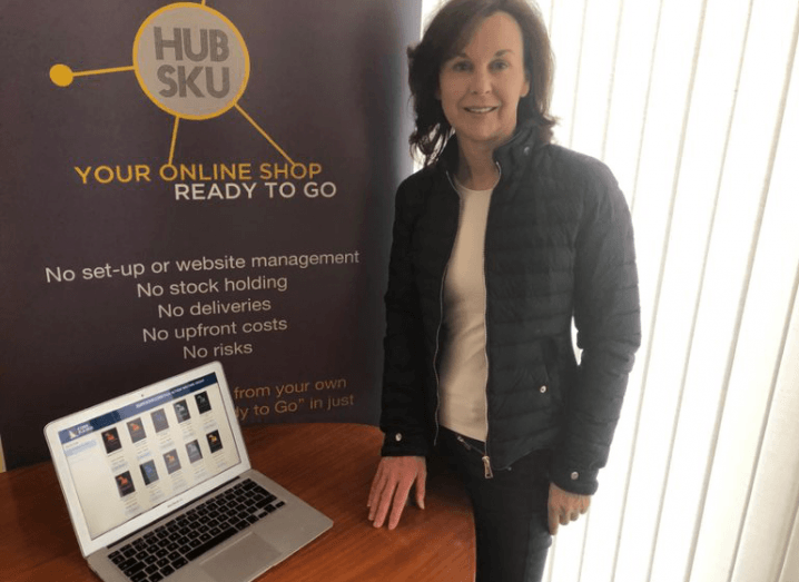 HubSku CEO Freda McEnroe stands in front of a MacBook on a table, beside a sign for her company. She has brown hair and is wearing a black quilted jacket.