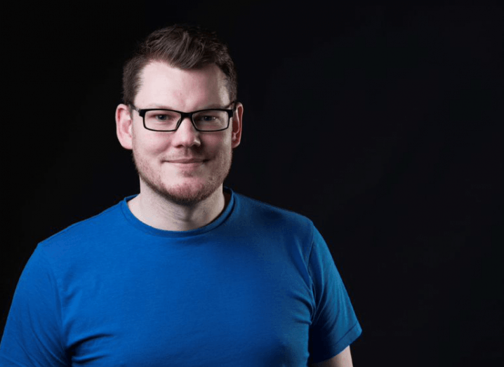 Brian D'Arcy standing in front of a black background, wearing a blue T-shirt and a pair of black glasses.