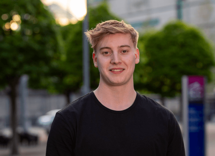 Young blonde man in a black T-shirt standing outside.