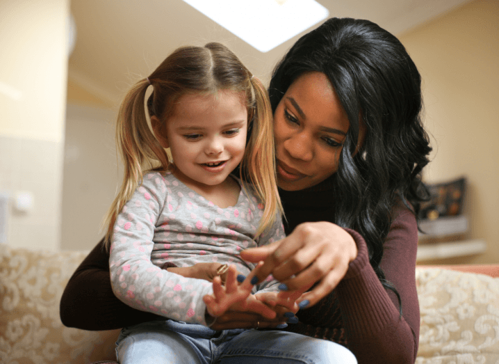 A woman helping a child count on her fingers. The woman has dark hair and is wearing a brown long-sleeved top, the child has blonde hair and is wearing a grey top with pink flowers on it.