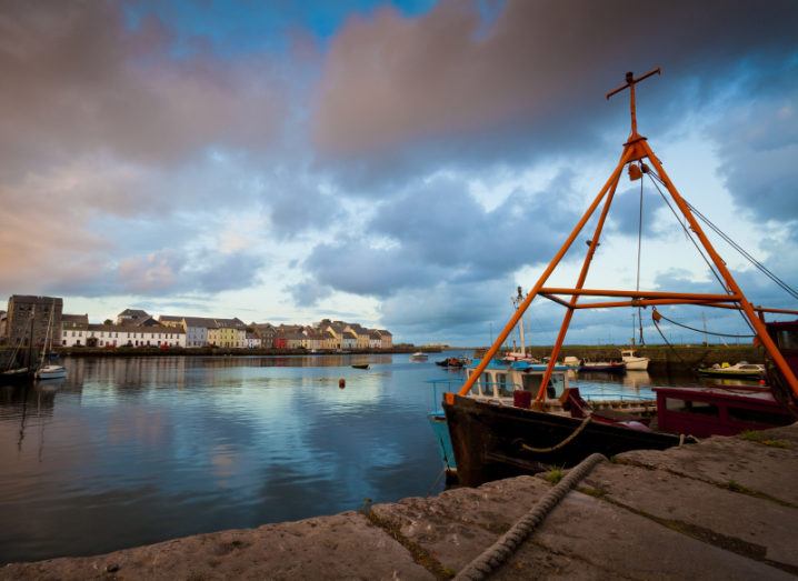 A view from the harbour of Galway city at sunset, with a boat in the foreground and the city’s colourful skyline in the background.