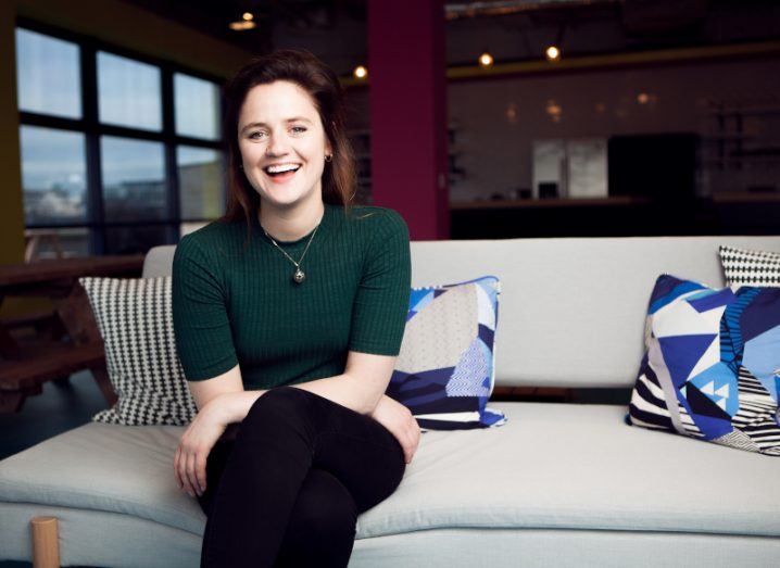 A young woman with dark hair and a green top sits on a sofa with blue cushions in a modern Huckletree office space.