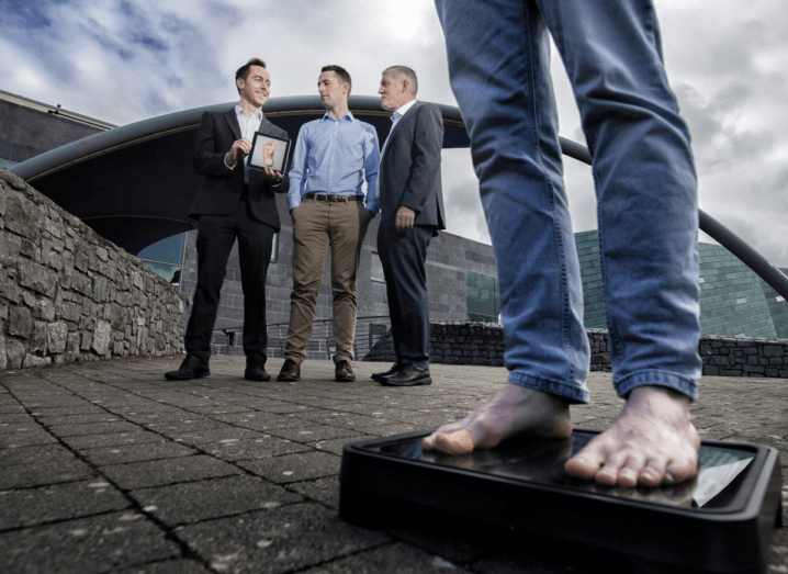 A person in blue skinny jeans stands on the Bluedrop Medical device while three men in the background hold a framed photo of a foot. They are all outdoors, in front of a tall grey building and a wall made of stone.