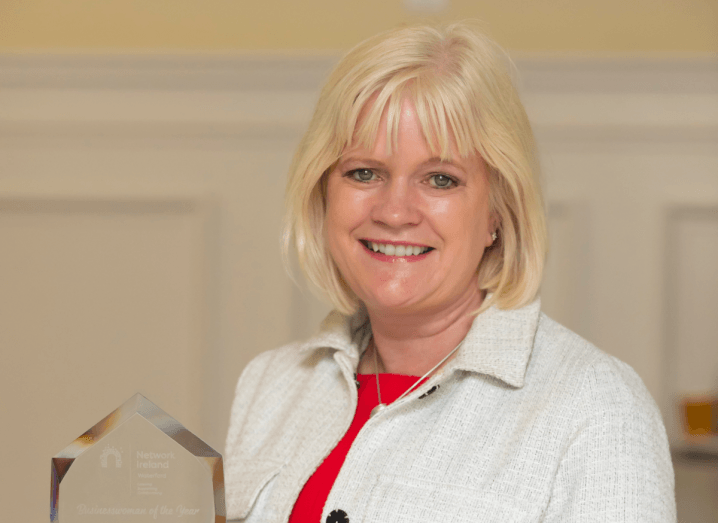 A blonde-haired woman with a full fringe smiles into the camera, holding a glass award. She is wearing a white jacket with a red dress underneath it.