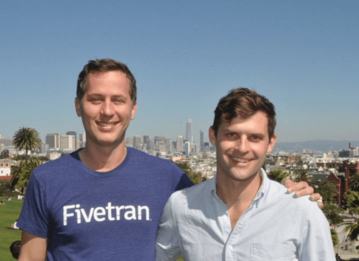 A man in a navy-blue t-shirt that says "Fivetran" stands beside a shorter man in a white shirt. They both have brown hair and are smiling. They are standing on a hill in front of the San Francisco skyline on a sunny day.