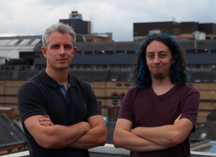 A man with grey hair stands with his arms crossed in a black t-shirt beside another man with long, black hair and blue streaks, who is wearing a burgundy t-shirt. In the background is Stephen's Green Shopping Centre's multi-story carpark against the Dublin skyline.