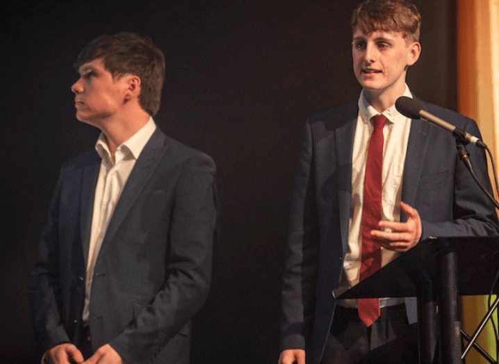 Two young men stand on a stage in dark suits and white shirts.