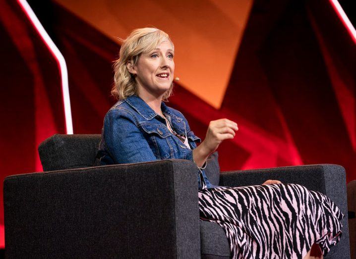 A woman seated on a stage with a red backdrop, wearing a head mic and speaking while gesturing with her hands.