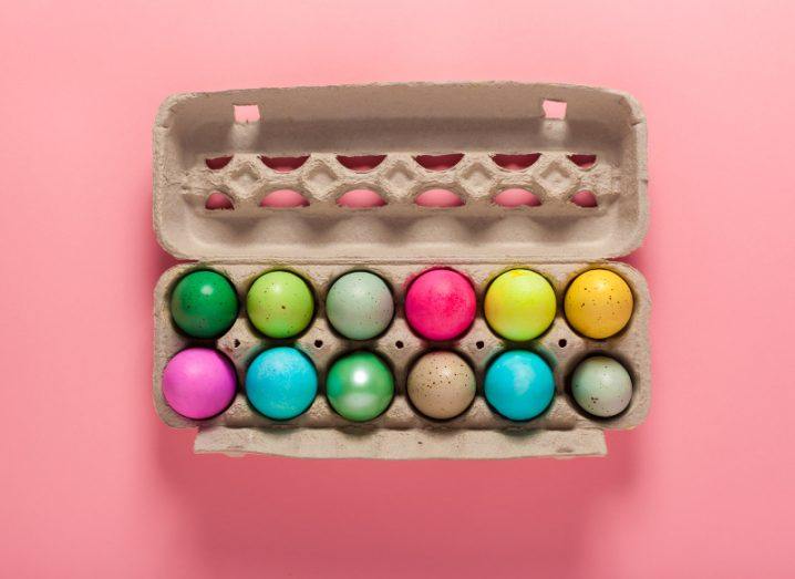 A batch of a dozen colourfully painted eggs in a carton on a soft pink background.