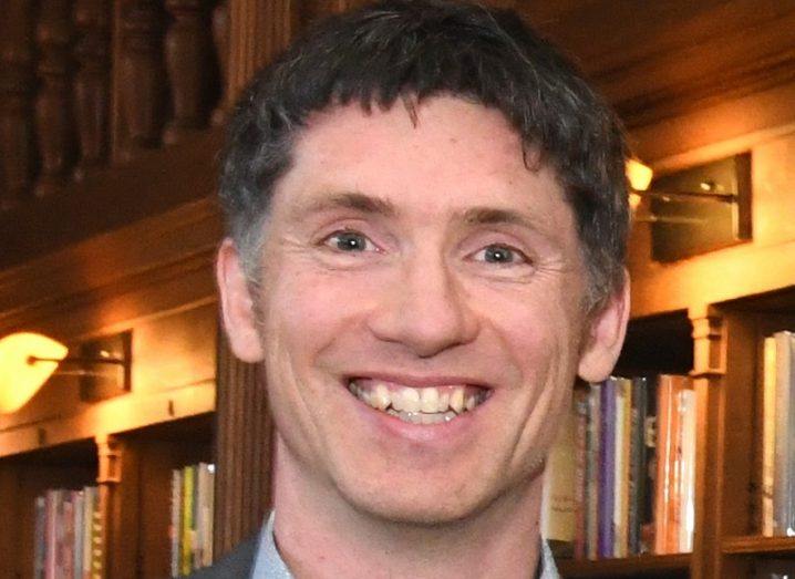 Headshot of Jamie Goggins smiling against a bookshelf background.