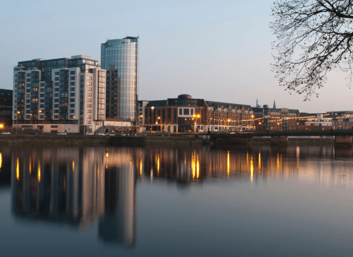 Tall buildings in front of a body of water in Limerick, at dusk. There are yellow lights in front of some of the buildings, which are reflected in the water.