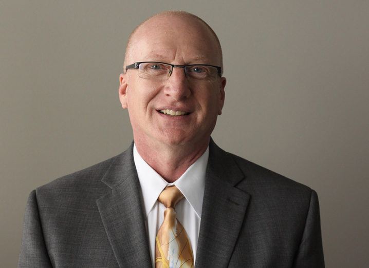 View of a bald man with glasses wearing a slate grey blazer, white shirt and gold tie.