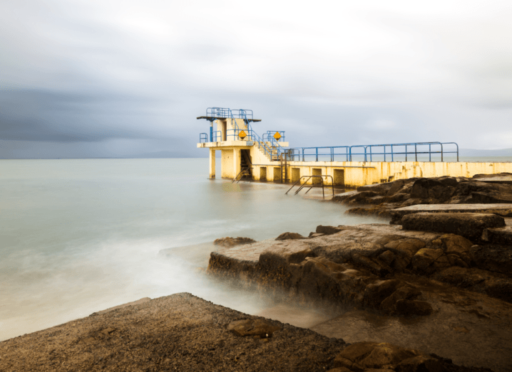 A yellow diving pier beside rocks and a body of water at Blackrock in Galway. There's a cloudy sky over the murky grey water.